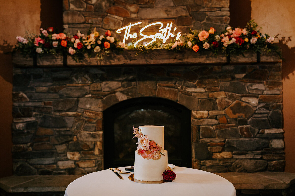 Wedding cake in front of fireplace with flowers and neon sign on the mantle at Aspen Lakes golf course in Sisters Oregon