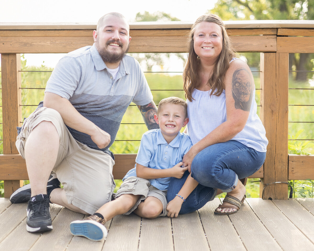 Mom, Dad, son sitting on a bridge at a wildlife sanctuary.  Dad is wearing a blue striped shirt, khaki shorts, black shoes, shaved head with a dark brown beard, smiling at the camera.  Young son has light brown hair, partial button up short sleeve blue short, khaki shorts, brown shoes, smiling at the camera.  Mom has long brown hair, wearing a sleeveless white shirt, blue jeans, and brown sandals, smiling at the camera
