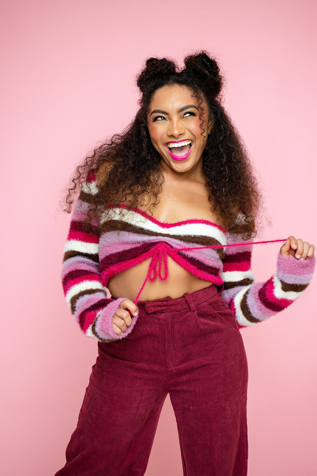 Fun and flirty portrait of a curly-haired teen pulling at her fuzzy striped crop top, standing in front of a soft pink background with an expressive smile.