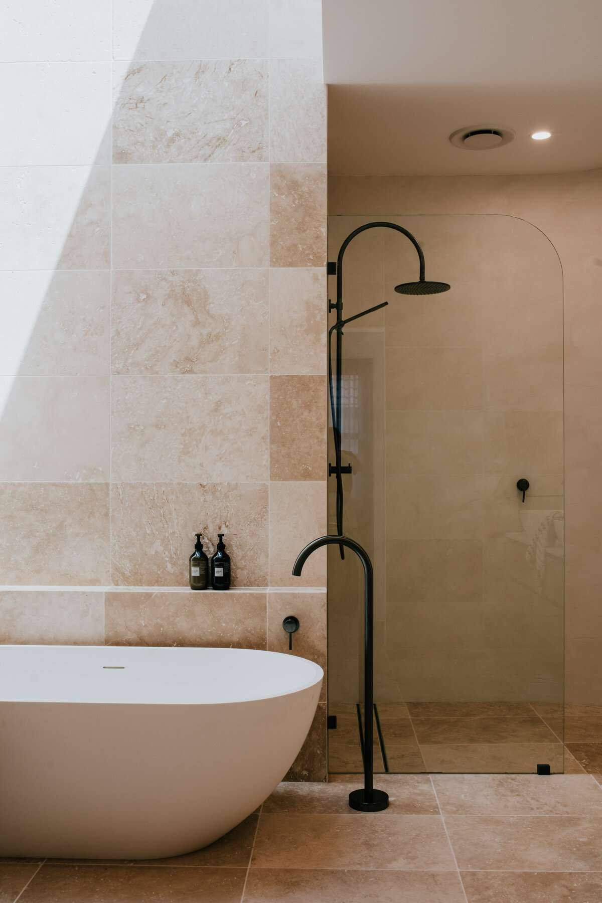 Minimalist bathroom designed with a white freestanding tub, black faucet, and beige stone tiles by a Newcastle Kitchen and Bathroom Designer.