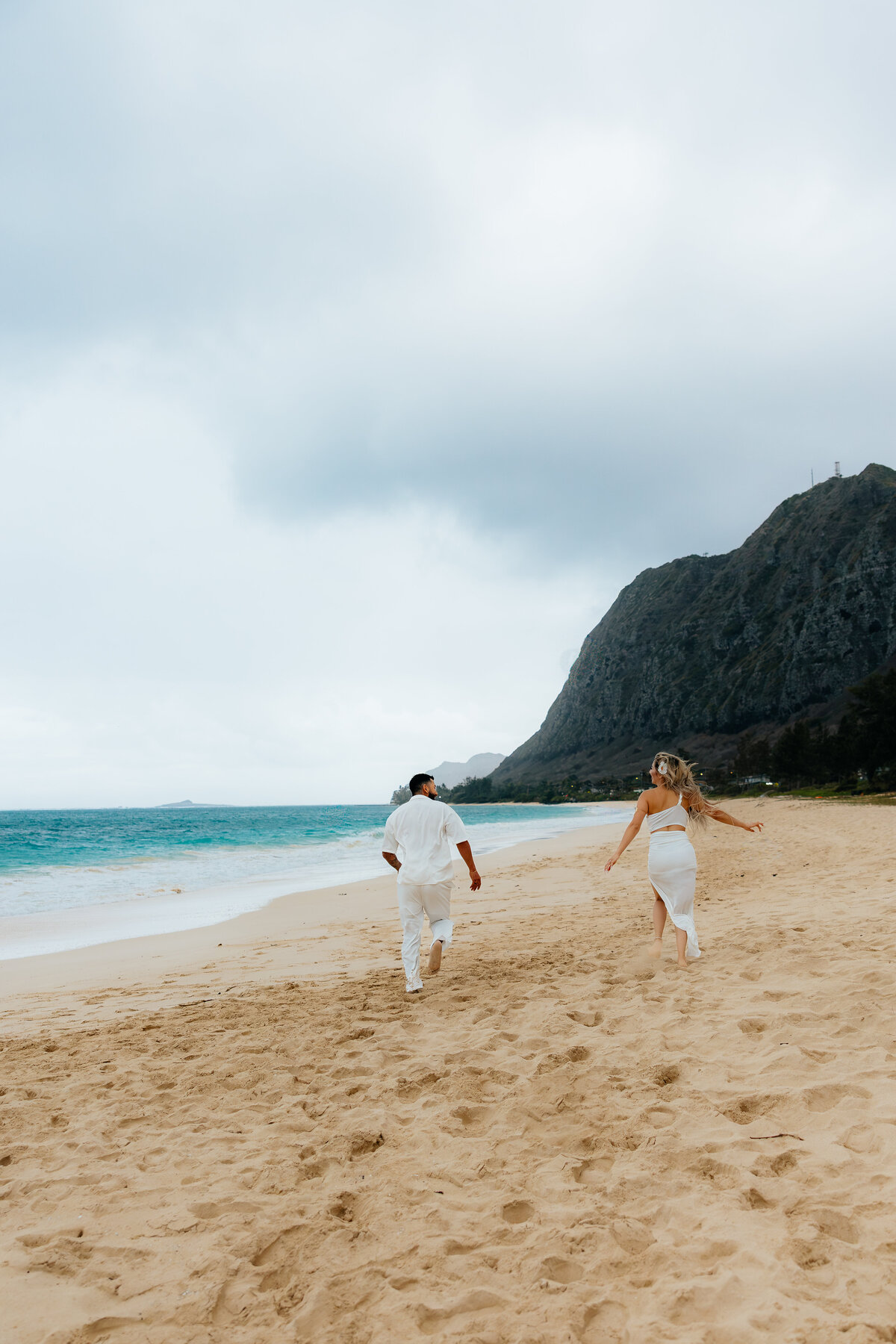 Couple in white outfits running along Waimanalo Beach with a  mountain backdrop on Oahu, Hawaii