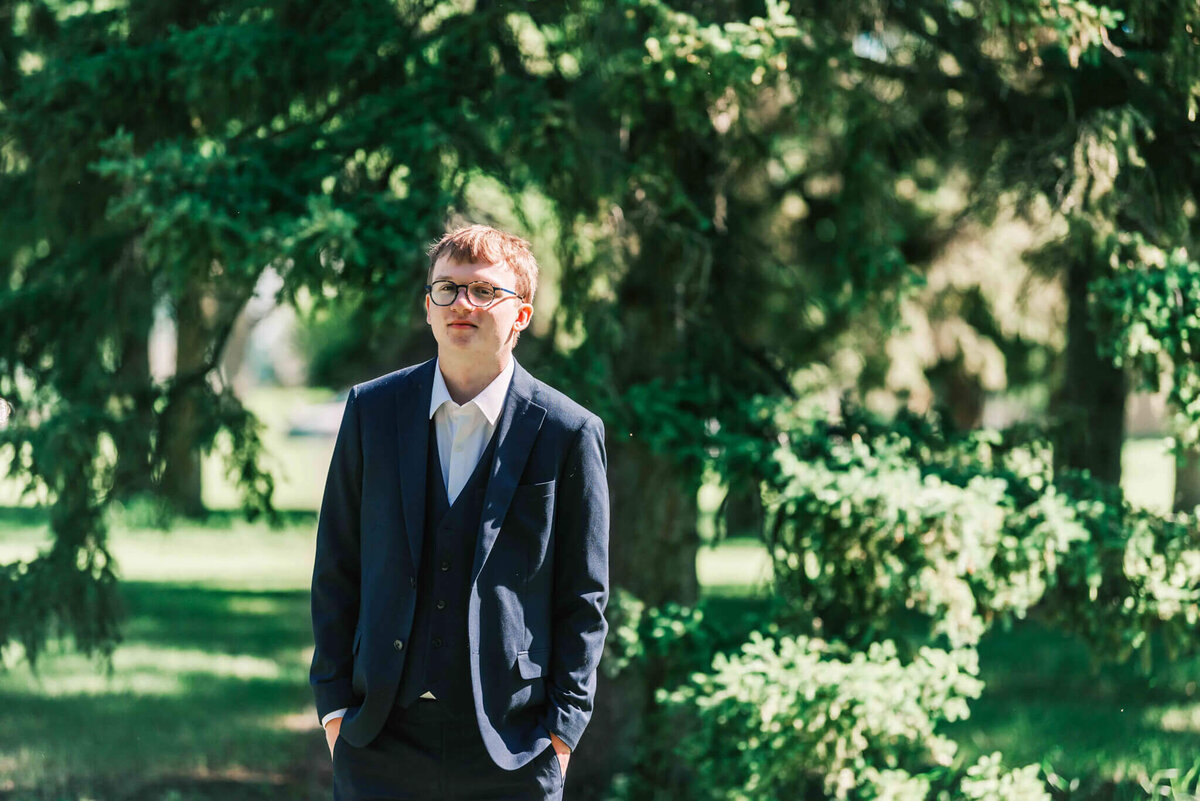 Young man stands with hands in his pockets in front of some trees. 
