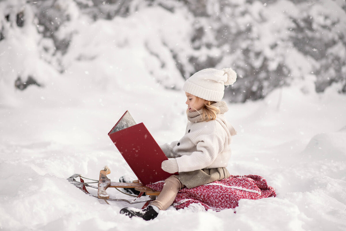 A little girl sits on a blanket on a wooden sled while reading a large red book.  It it winter and lots of snow in Kamloops. Portrait by Brittany Danielle.