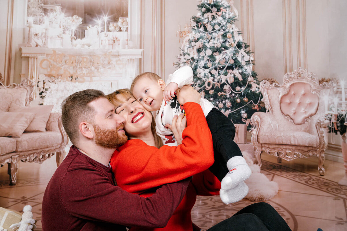 Parents cuddling their baby during Christmas photo session in Calini Weddings Studio - elegant festive tree and warm tones.