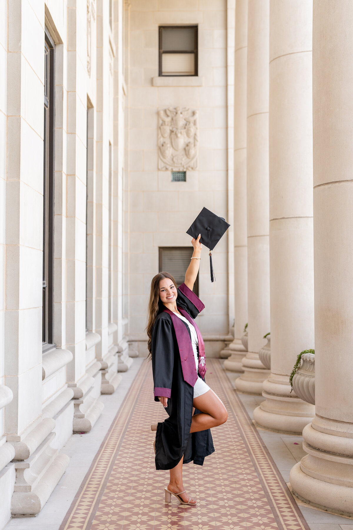 Texas A&M senior girl  wearing gown and stole and holding up cap celebrating and smiling within the columns of the Administration Building