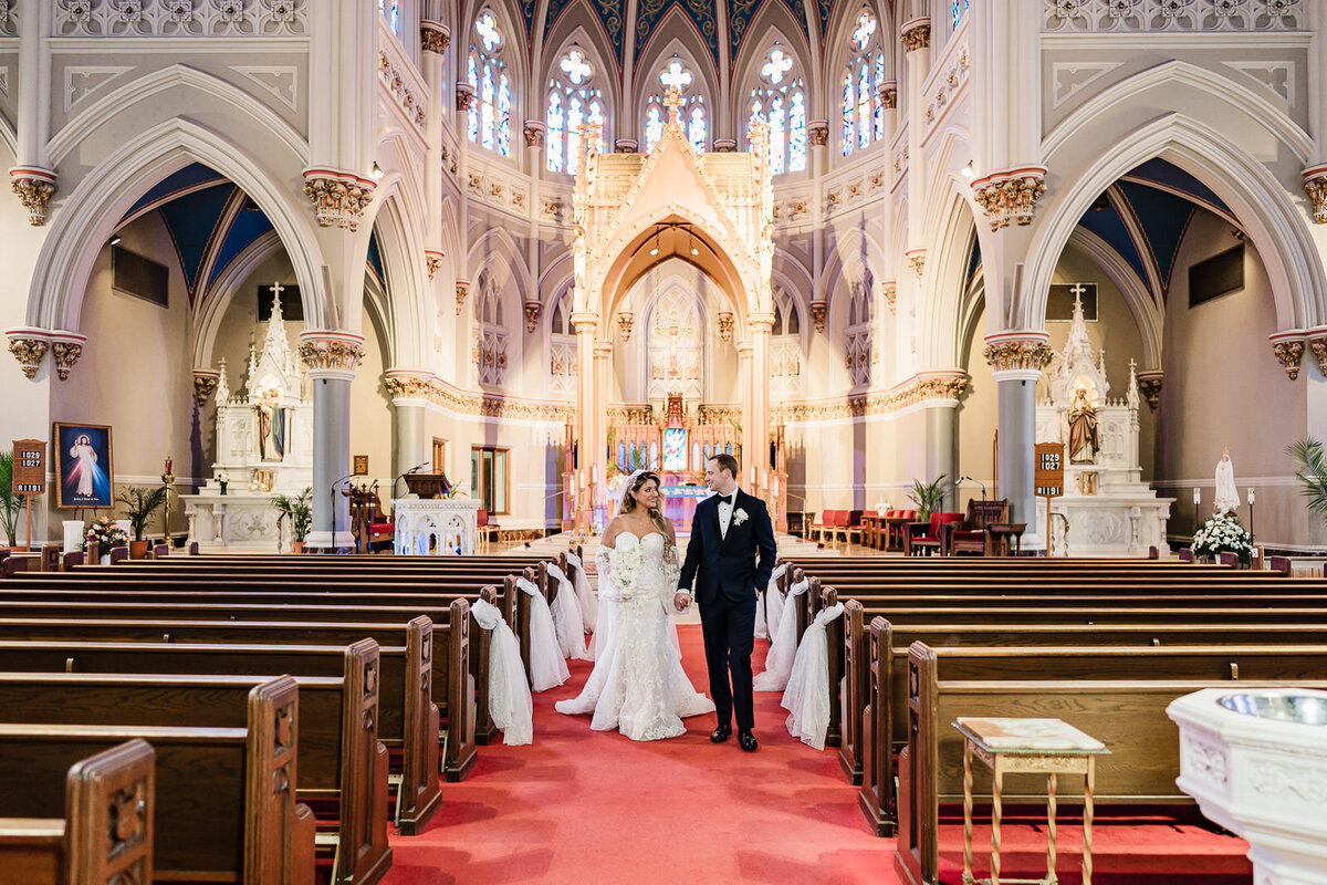 Bride and groom walking hand in hand down the aisle of a grand cathedral, smiling at each other with ornate arches and stained-glass windows behind them.