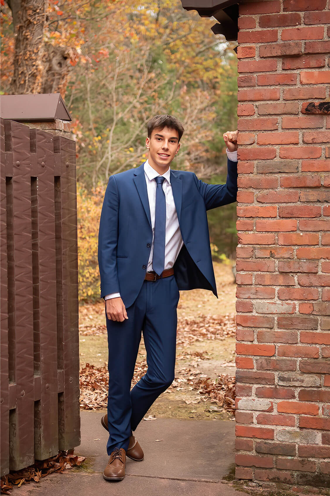 A senior in a navy suit leans with one arm against a brick wall at Whip Hill Park in Stoneham.