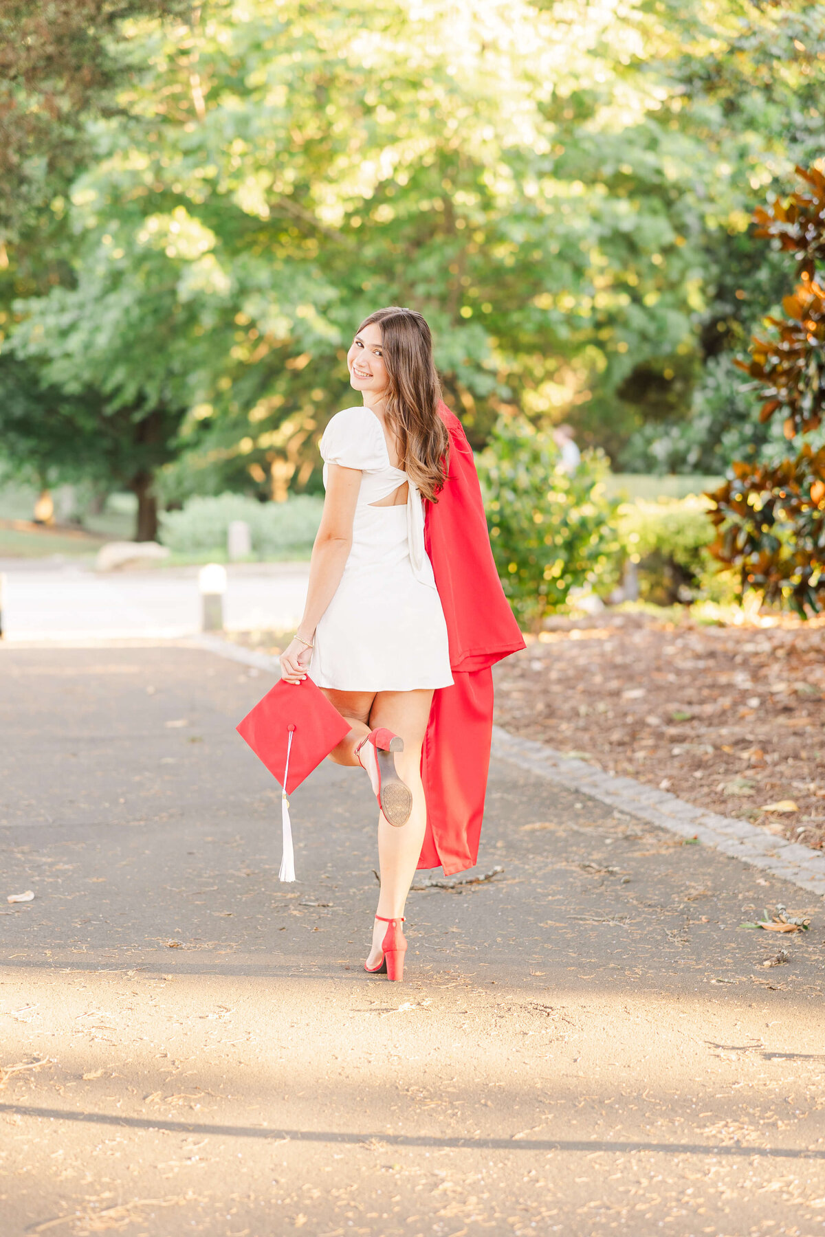 A young woman strolls along a sunlit path surrounded by greenery in Fayetteville, NC. Clad in a white dress and red stole, she clutches a red graduation cap with a tassel. Her red high heels complement her look as she glances back, smiling for the senior portrait photographer.