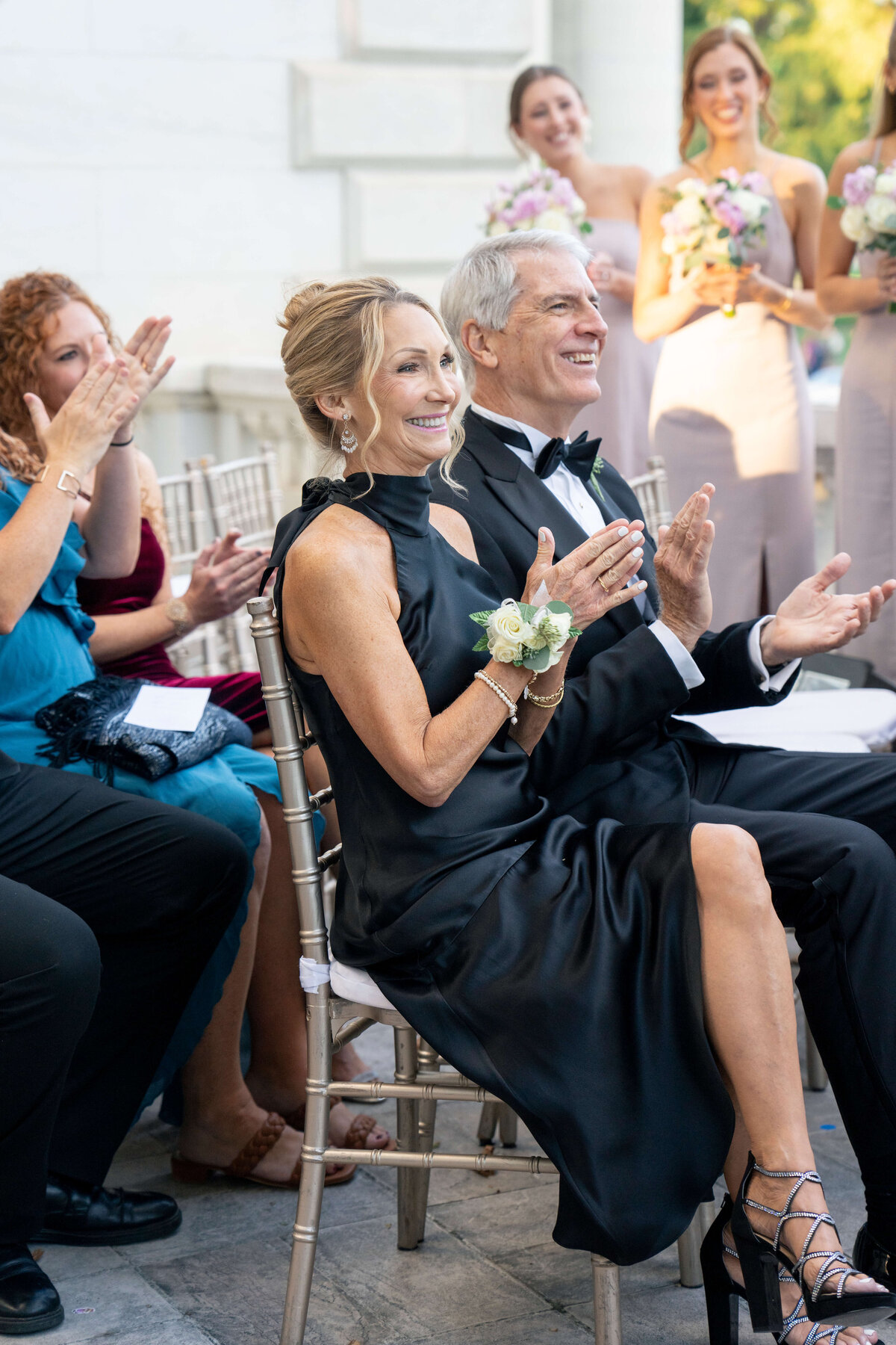 A couple, dressed elegantly for an event, sits among others applauding happily. The woman wears a black dress and the man is in a suit with a bow tie. People around them are smiling and holding bouquets, suggesting a celebratory occasion, possibly a wedding.