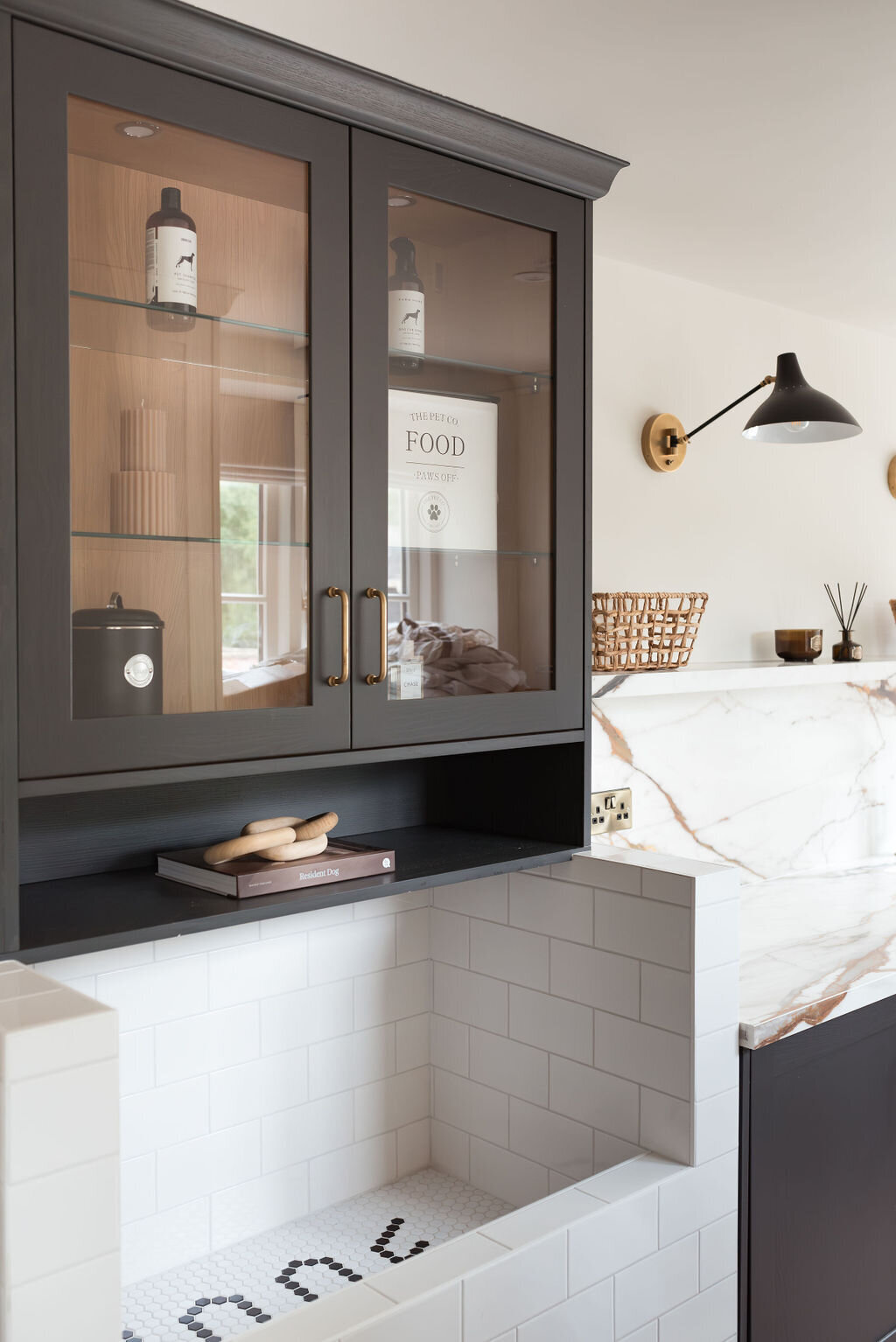 A modern dog wash room with black cabinets, a large white subway tiled washing station and white marble countertops. A black built-in shelf sits above the washing station. A shelf with wicker baskets sits above the counter, along with two black lights.