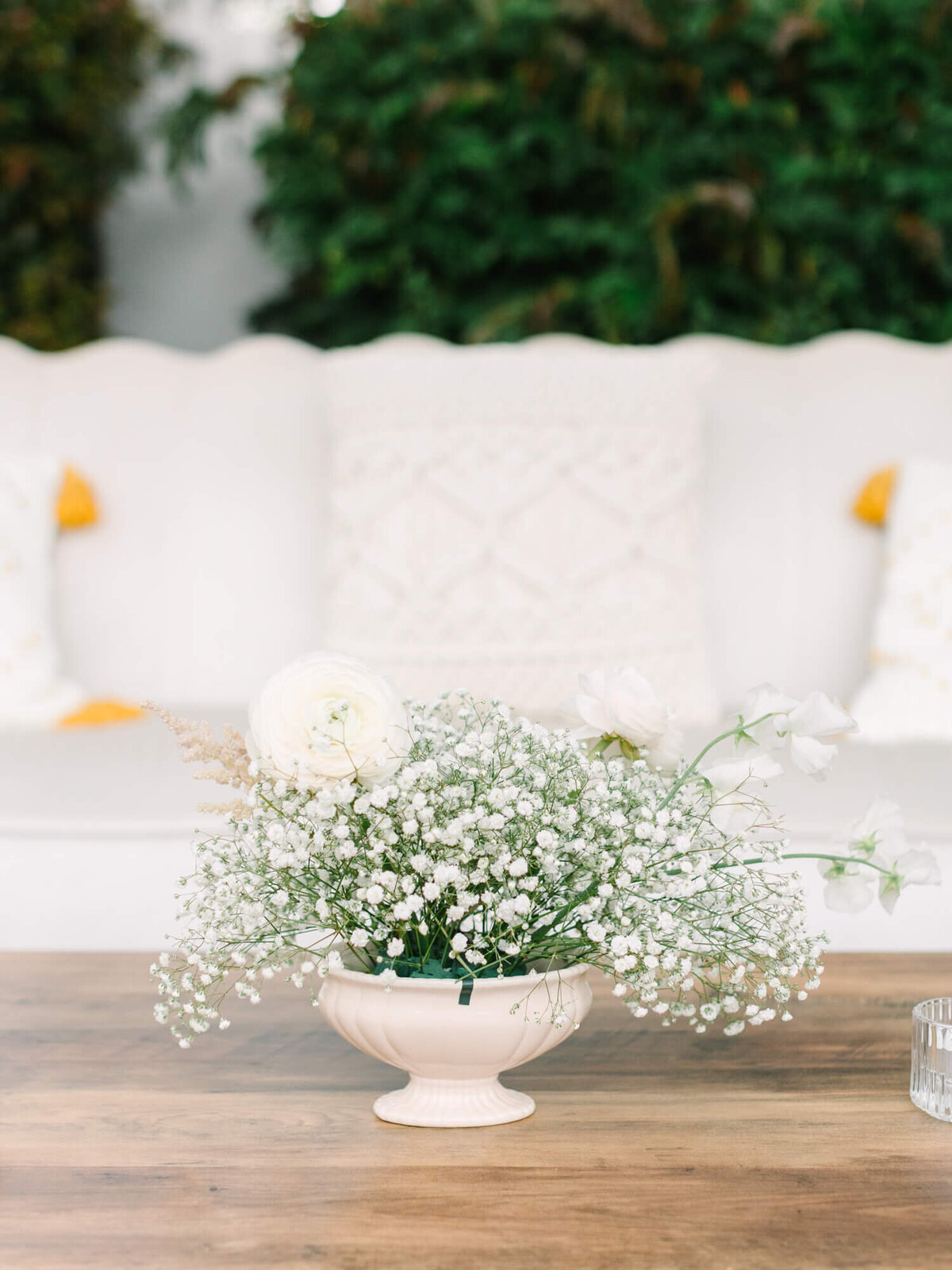 A serene floral arrangement with white roses and baby's breath in a cream vase on a wooden table. Soft-focus couch and greenery create a tranquil backdrop.