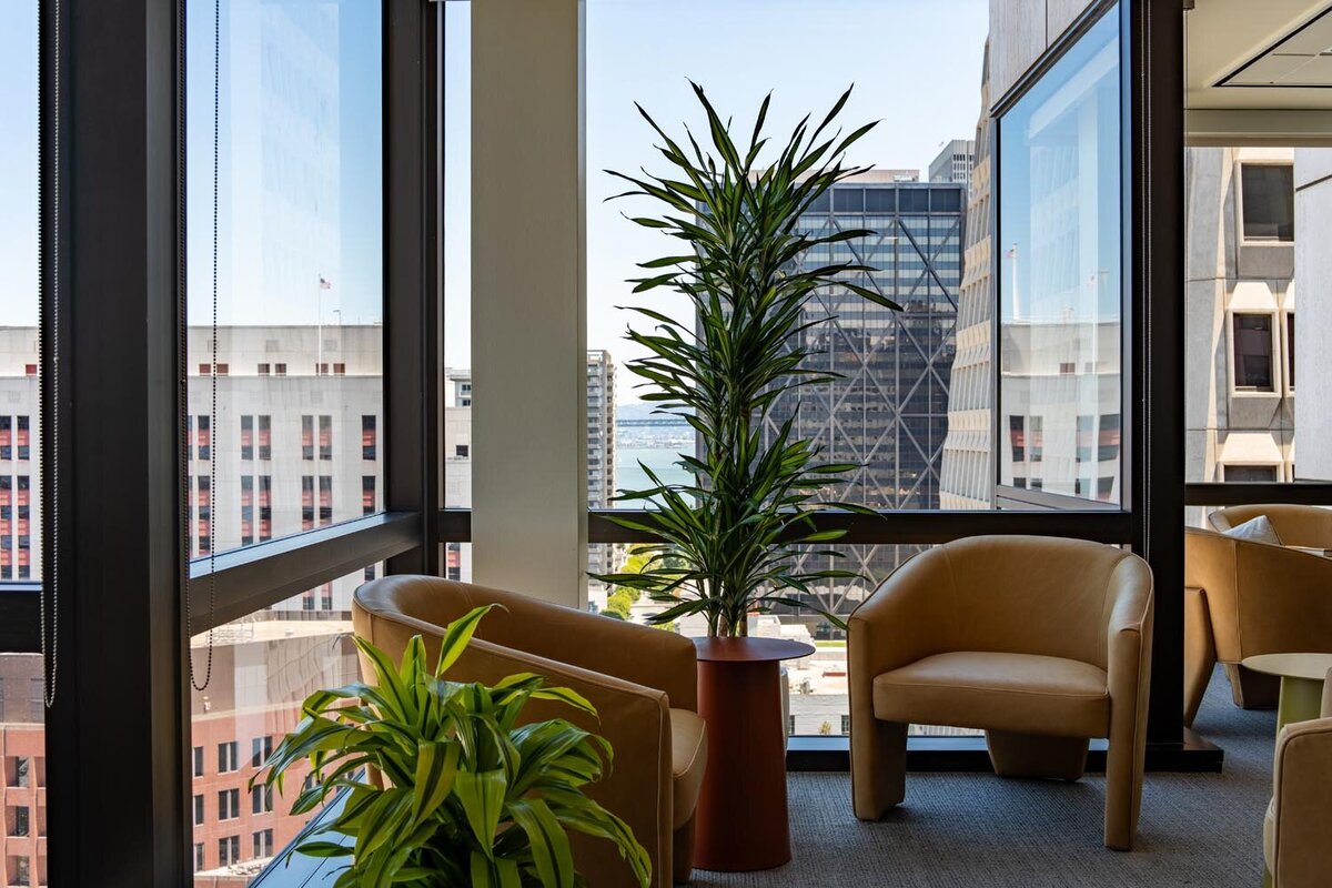 Corner lounge area with tan chairs, potted plants, and floor-to-ceiling windows showing downtown city views.