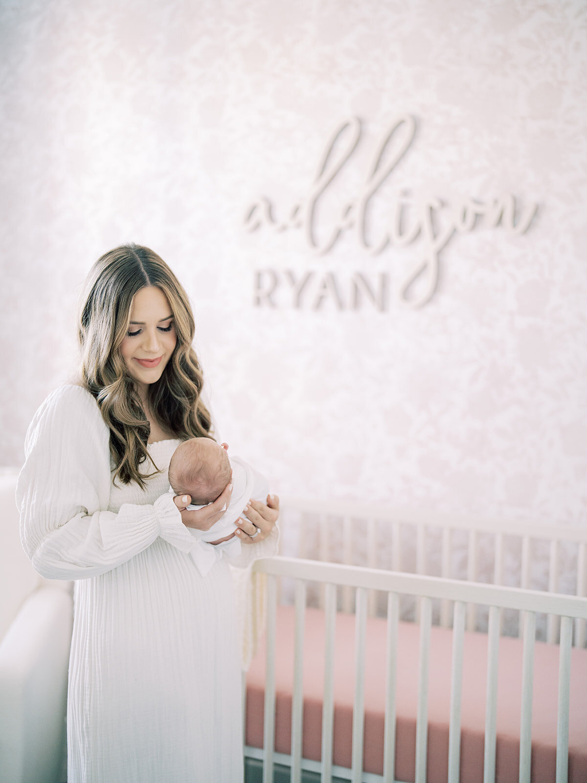 Mother with long brown hair in Nothing Fits But dress holds her newborn baby girl in her pink nursery.