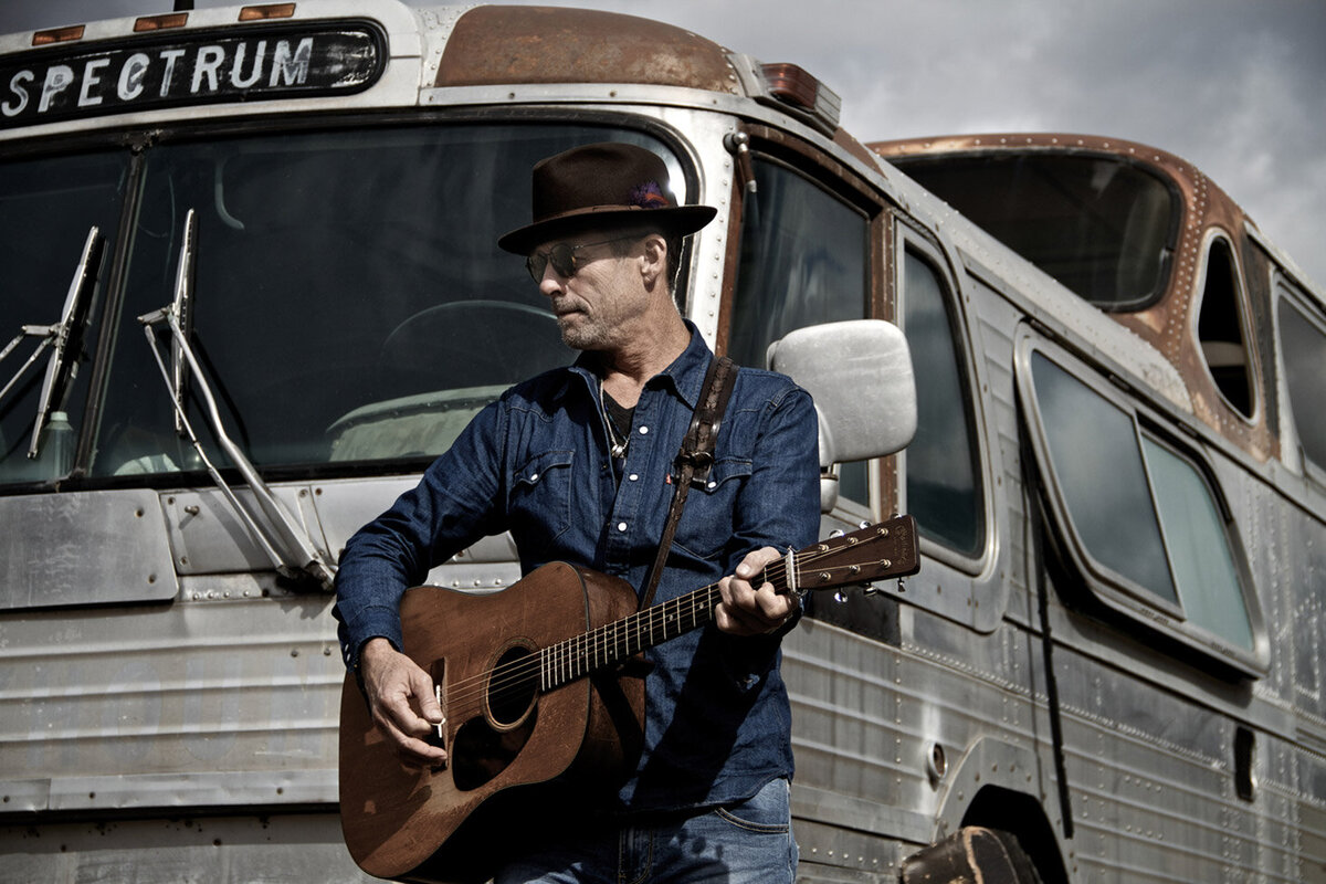 Musician Portrait Barney Bentall standing in front of vintage metal bus holding brown acoustic guitar Roadshow Gallery