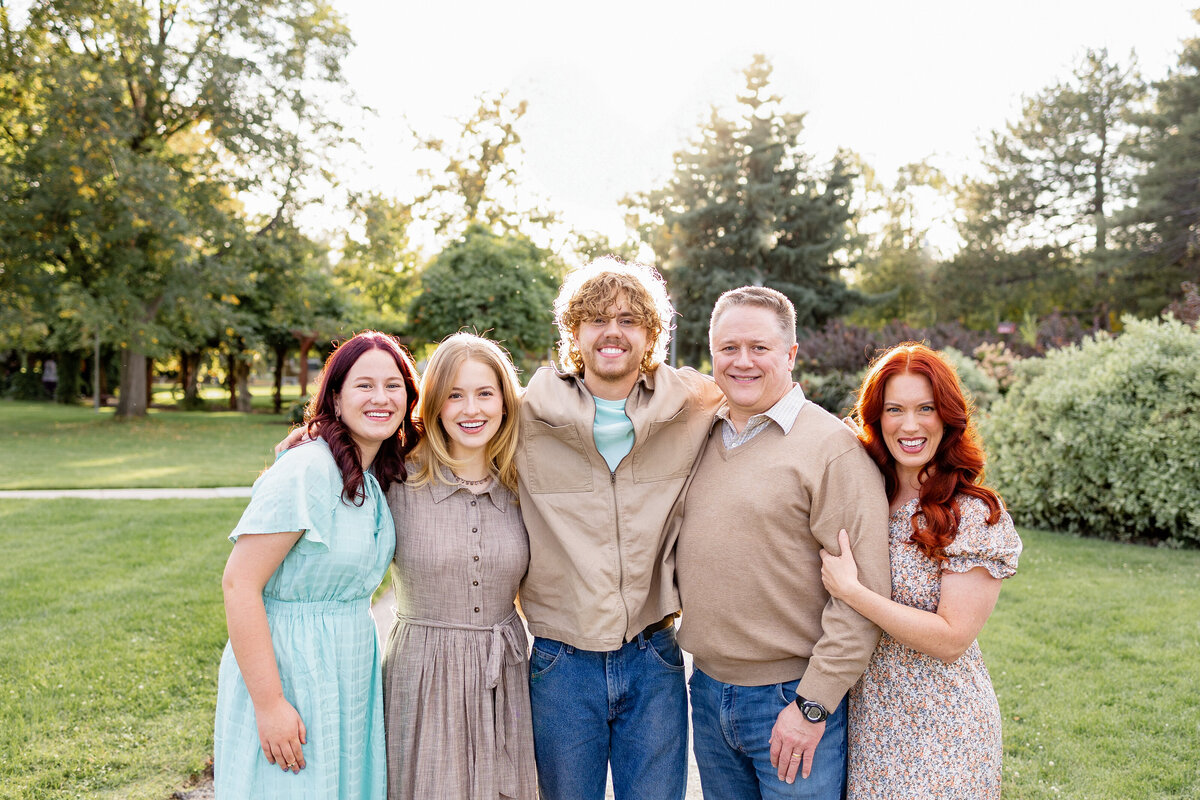 A family of five stands together on a garden pathway, illuminated by warm evening sunlight with tall trees in the background.