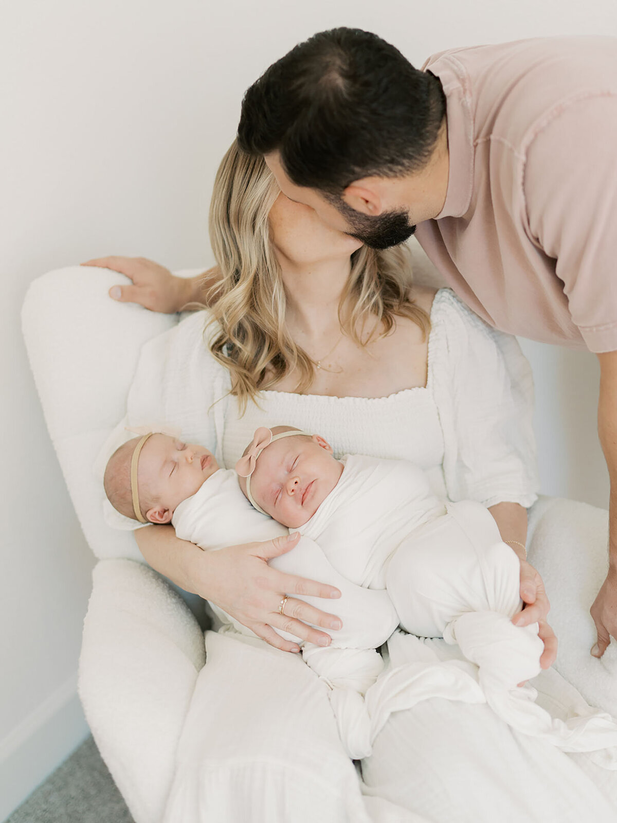 A heartfelt moment from a newborn session featuring a mother gently cradling her baby while the father leans over to kiss her. The soft lighting and warm tones highlight the love and connection between the new parents and their little one, creating a tender and intimate scene.