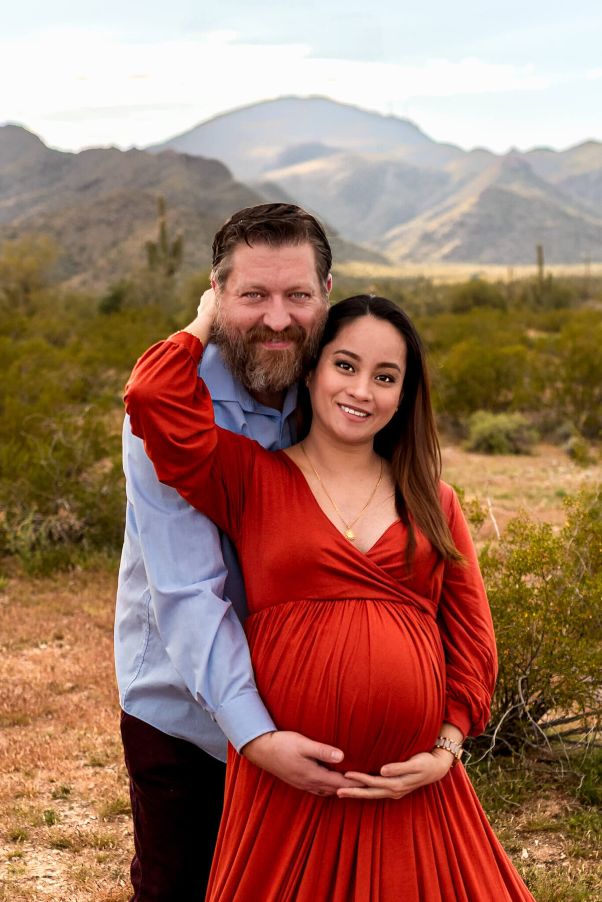 Expecting mom touching husband while he stands behind her in a hug. Arizona maternity session by Cactus & Pine Photography