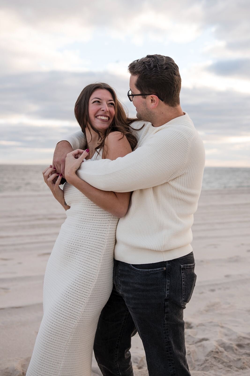 Pcture of young enggaged couple wearing off-white knitted garments as they hug.