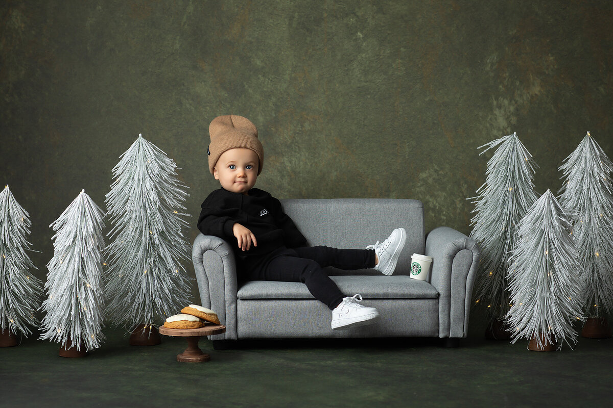 A stylish toddler lounges on a mini gray couch, dressed in a black outfit and tan beanie, surrounded by frosted Christmas trees, cookies, and a mini cup of Starbucks. This fun seasonal setup was captured by a Dallas milestone photographer.