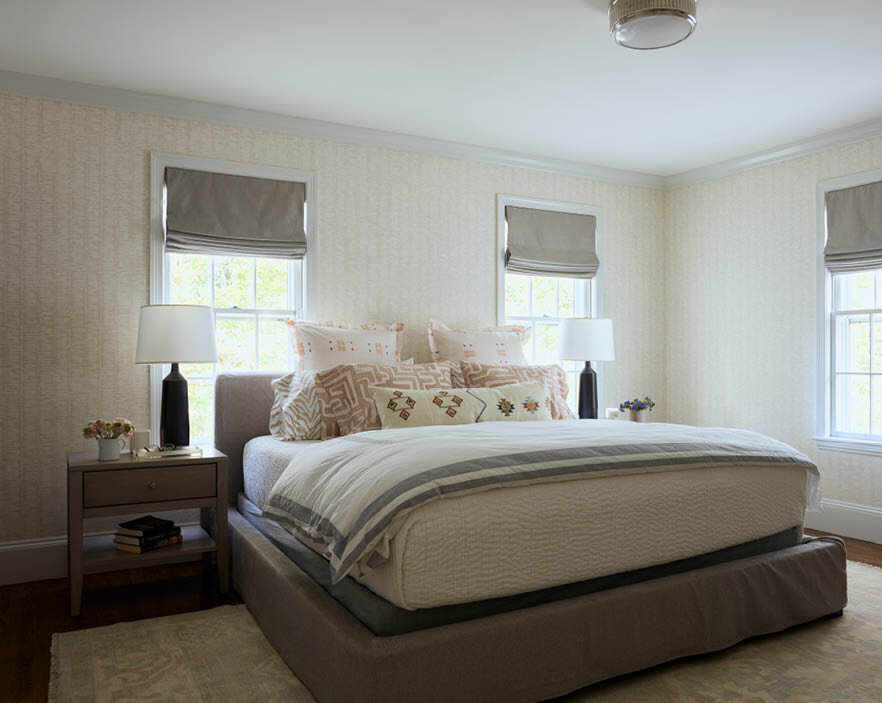 Explore the primary bedroom in a historic Hingham home, featuring tone-on-tone wallpaper, neutral trim, light grey linen window treatments, an upholstered bed, and minimal decor on bedside tables.