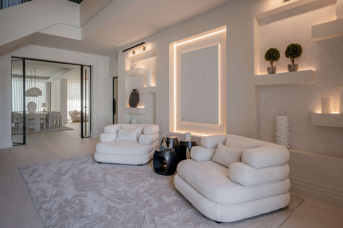 A spacious, modern entry area at the foot of the stairs, with a focus on light, neutral tones. There are two plush, light-colored armchairs and a soft, textured rug. Built-in shelves with decorative items and a wall with the illuminated text 'FOREVER ISN'T LONG ENOUGH' add a sleek coolness to the space. In the background, a dining area with a large table and pendant lights is visible through glass doors.