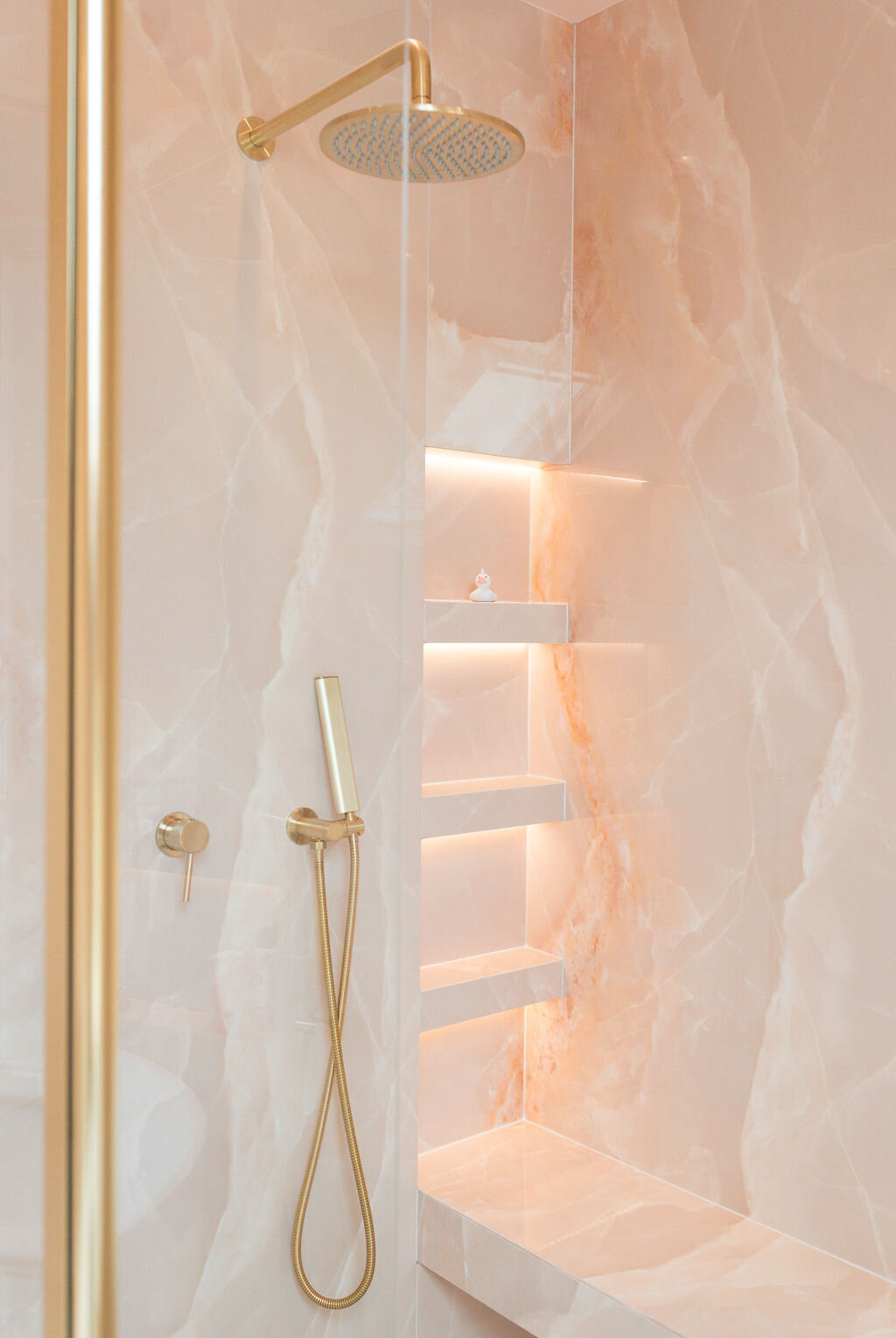 A luxurious glass-enclosed walk-in shower, featuring pink marble walls, gold hardware, built-in shelving, and a bench.