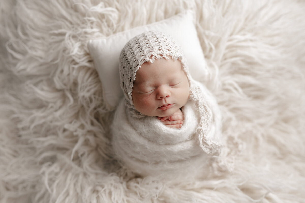 Baby girl swaddled for newborn portraits in knit wrap and crochet bonnet. Baby's hands are peeking out of the wrap and folded under her chin. Baby is laying on a matching cream flokati with newborn sized pillow resting under her head.