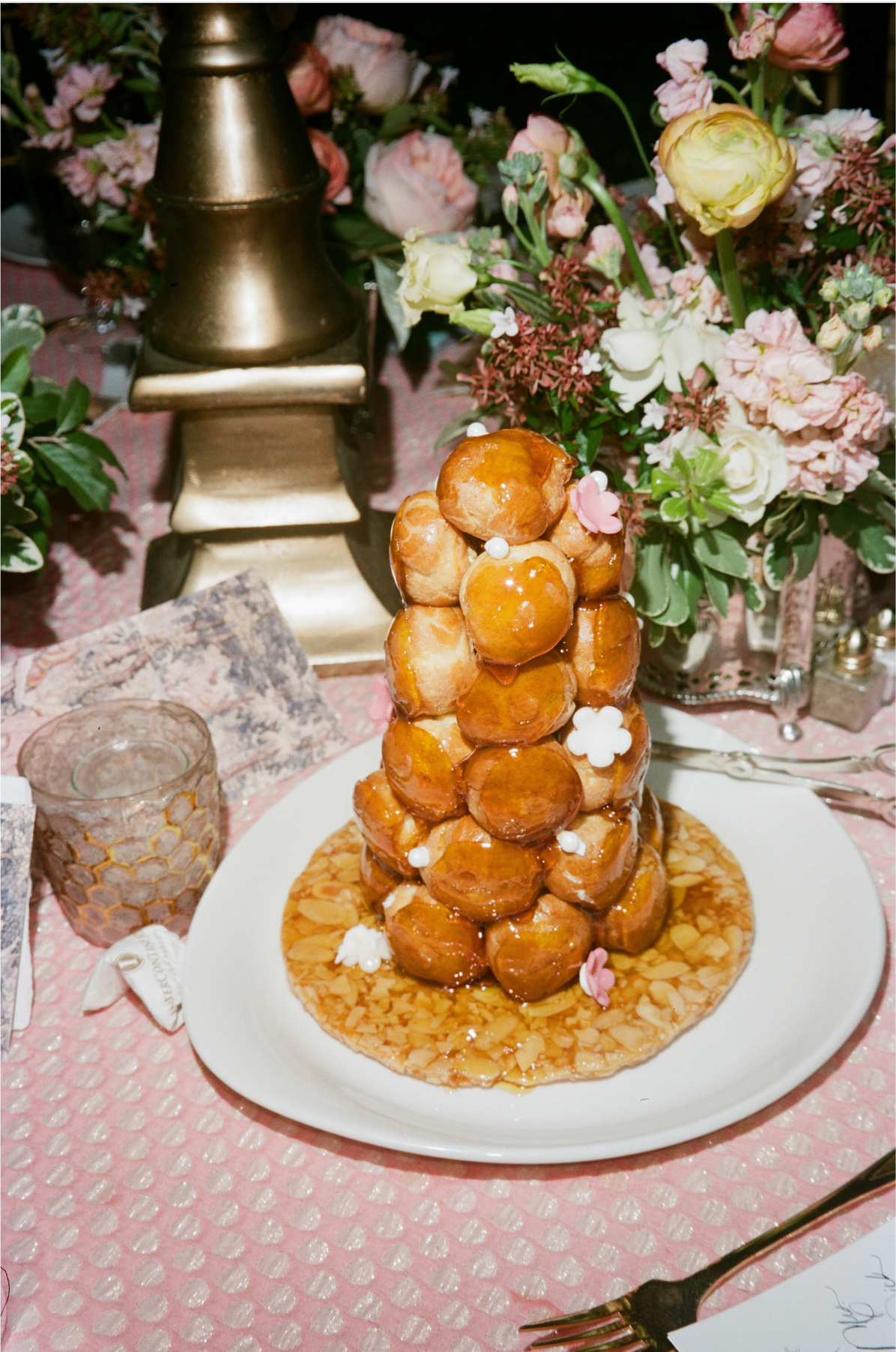 The photo was taken at The Willard Intercontinental in Washington D.C., known as one of the top wedding venues and hotels in DC. Captured by photographer Shawn Connell, the scene features a stunning display of a croquembouche, a traditional French dessert. The tower of cream-filled profiteroles is artfully stacked into a cone shape, garnished with delicate small flowers. It sits elegantly on a white plate on a table adorned with a pink tablecloth, beautiful floral arrangements, and a shimmering gold lamp, creating a truly enchanting setting.