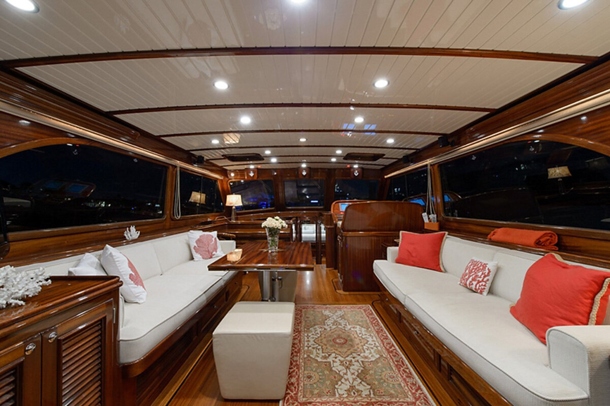 Elegant yacht interior with wooden paneling and ceiling, white cushioned seating adorned with red and white pillows, a patterned rug in the center, a small table with flowers, and large windows on both sides offering views of the night sky.