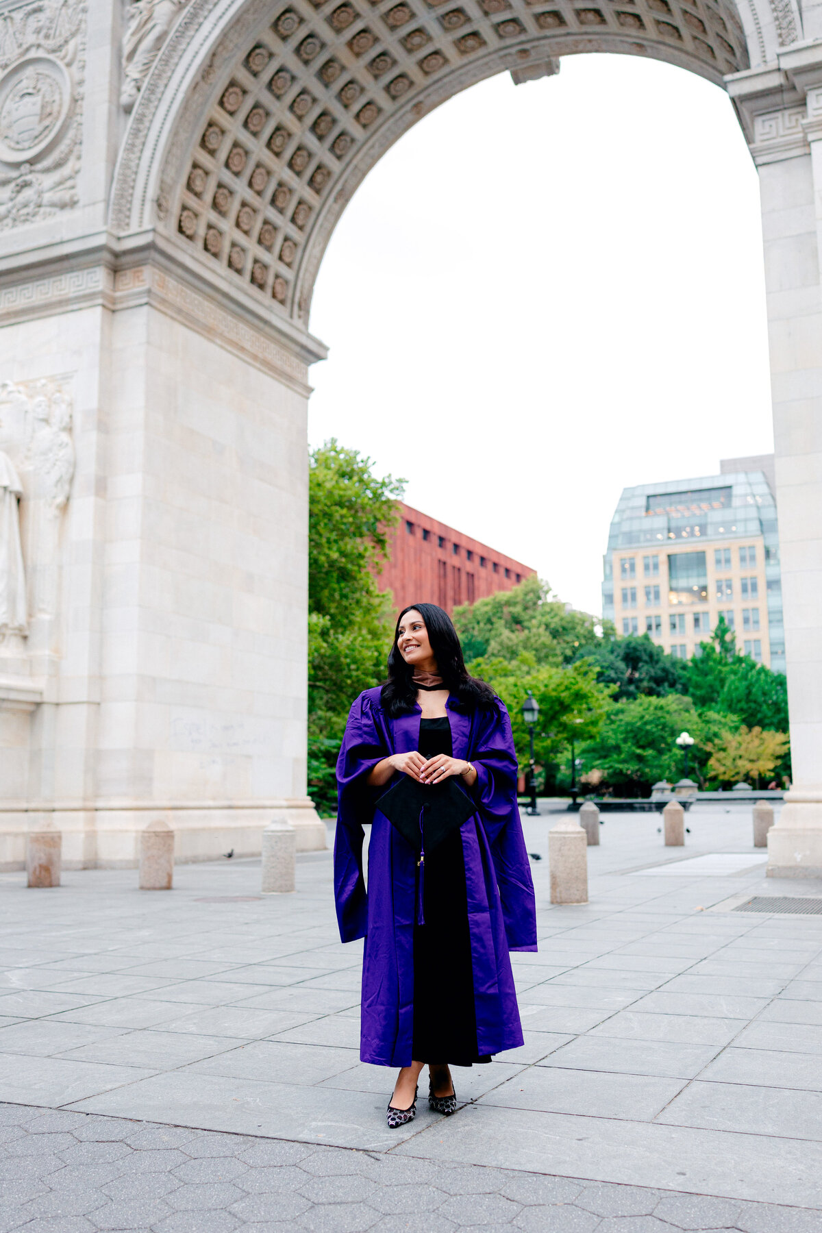 NYU graduate smiling in purple gown in front of the Washington Square Park arch.