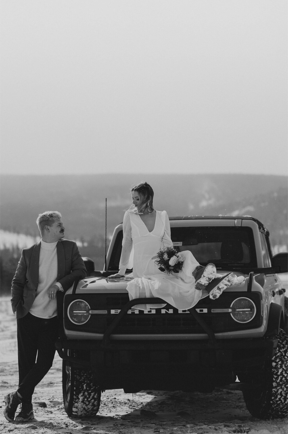 bride sits on the hood of her jeep bronco while she holds flowers and looks at the groom during their elopement on a snowy day in Bend, OR