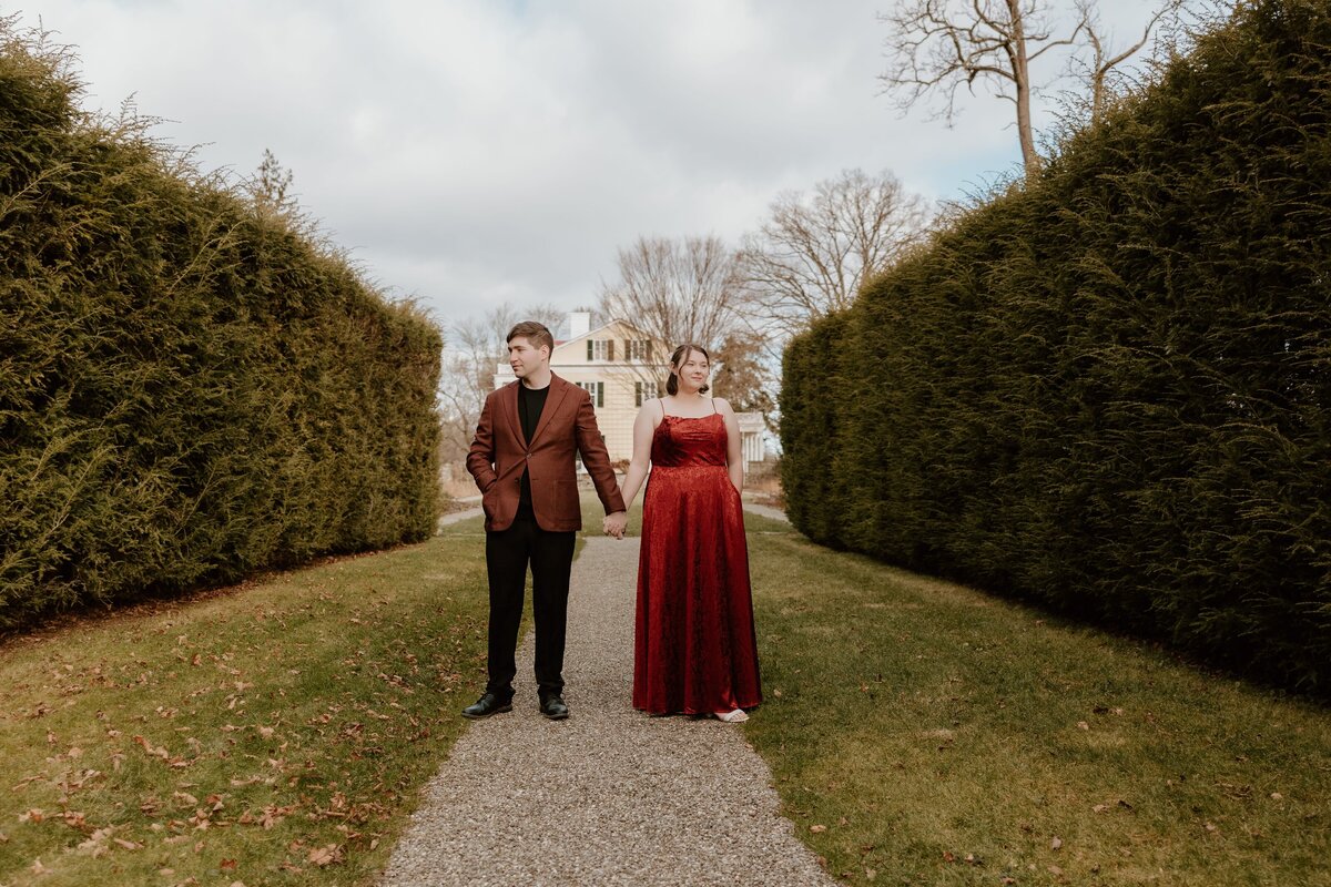 Romantic couple holding hands on a tree-lined pathway at FDR Historic Site in Hyde Park, wearing a red formal dress and brown blazer.