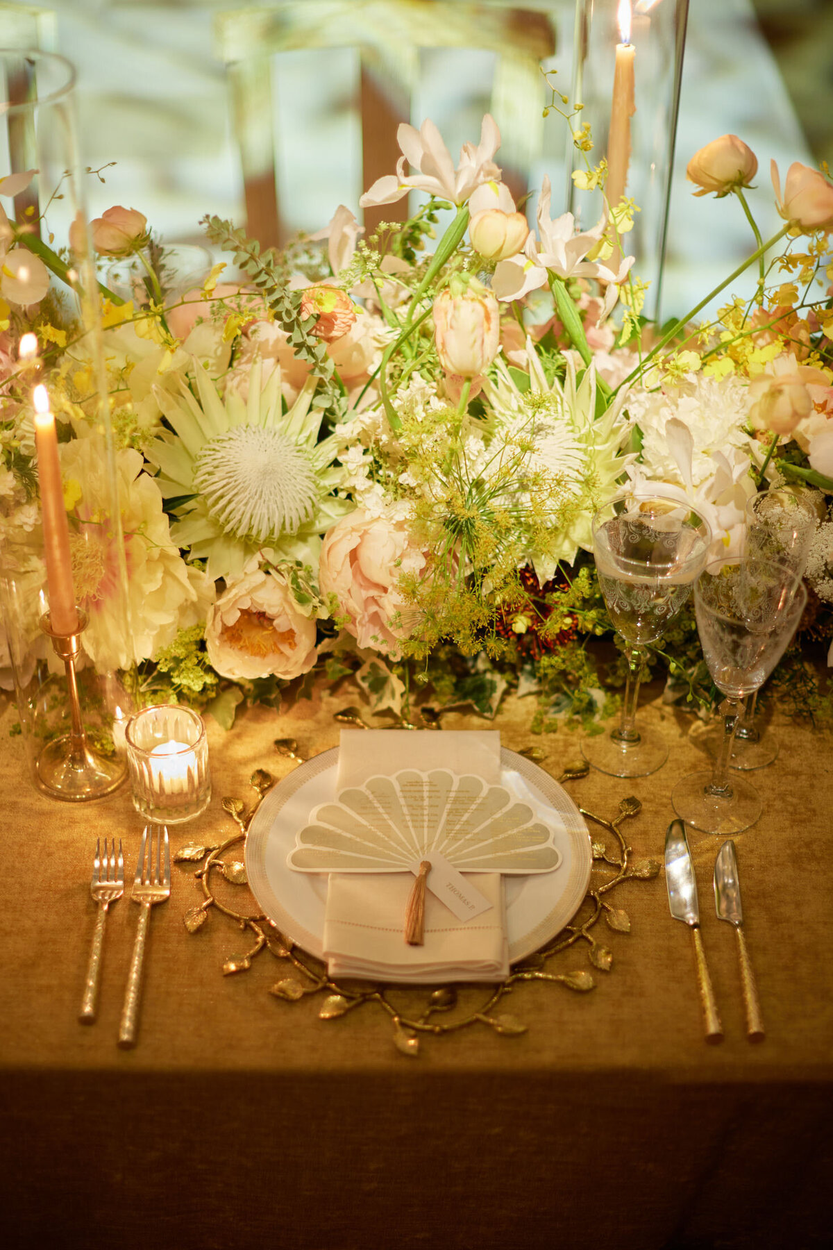 The photo was taken at the New York Public Library in New York City by photographer Shawn Connell. It features a beautifully arranged table setting with a botanical theme. The centerpiece showcases lush flowers, including roses and chrysanthemums. A white plate with a napkin in the shape of a seashell, surrounded by elegant cutlery, sits atop a gold tablecloth. Candles add a warm glow to the overall ambiance. Videography by Aaron Navak Films.