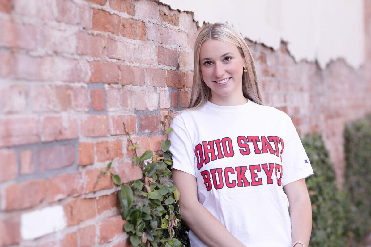 A person with long hair wearing an Ohio State Buckeyes t-shirt stands against a brick wall with greenery. They are smiling and the wall shows some wear and tear.