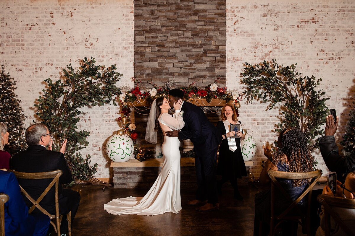 The bride and groom kiss in front of the decorated mantle at Steel Magnolia Barn. Their christmas wedding is decorated with oversized ornaments and a greenery arbor against a white washed brick wall. The groom is wearing a black suit and the bride is wearing a fitted satin gown with long sheer sleeves and a fingertip veil.