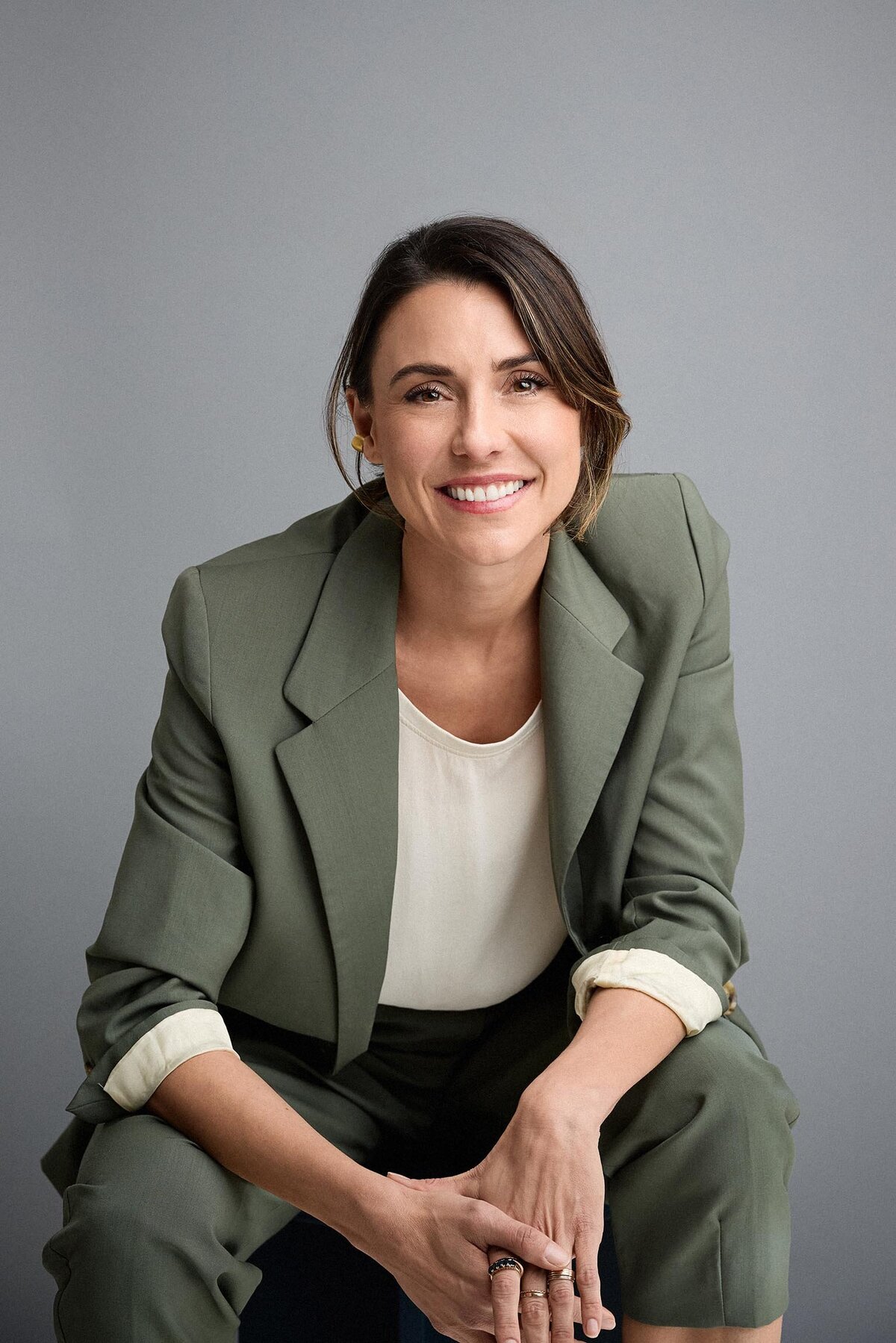 Confident woman in sage suit and cream top against a grey studio background. Sitting looking directly at camera.