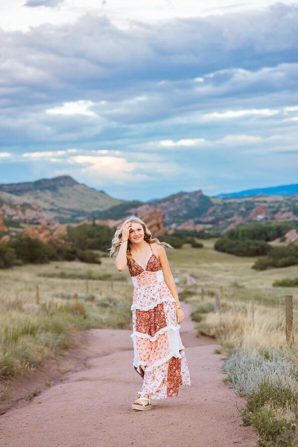 Girl fixes hair while walking on trail with mountains behind her.