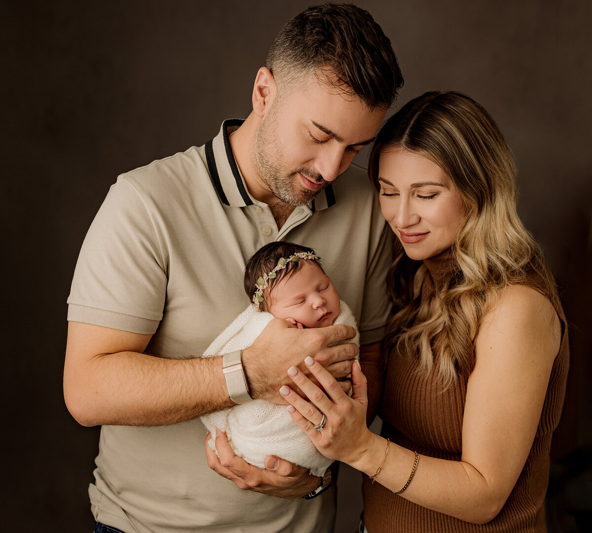 dad in a grey beige shirt and mom in a brown halter top holding newborn abby girl wrapped in a cream swaddle and wearing a leaf headband on a dark brown backdrop 