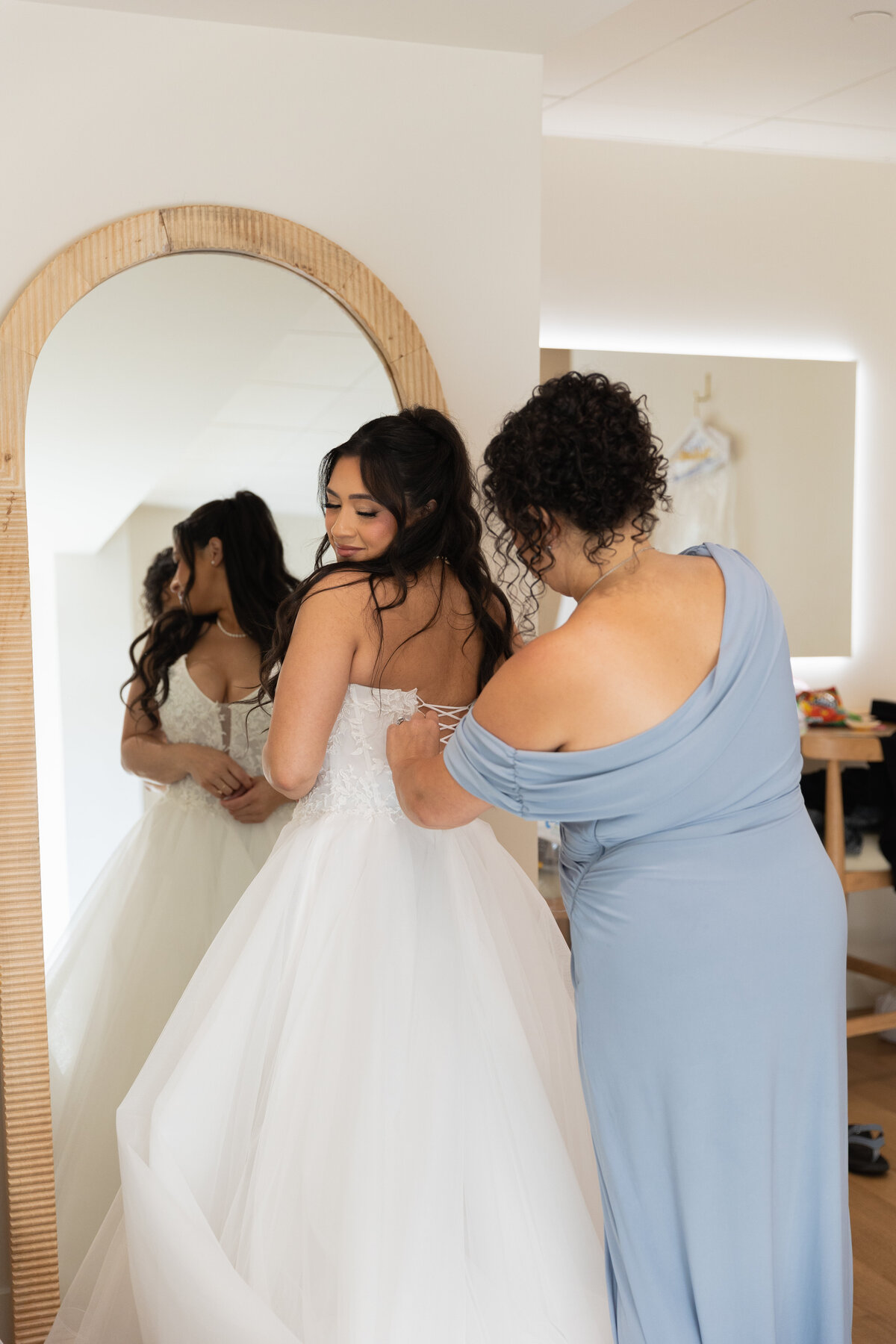 The Lakehouse Inn MA  | Kelsey Sheehan Photography Timeless Rhode Island Weddings | A bride in a white gown stands before a mirror while a woman in a light blue dress helps adjust her dress. The room is softly lit, conveying warmth and anticipation.