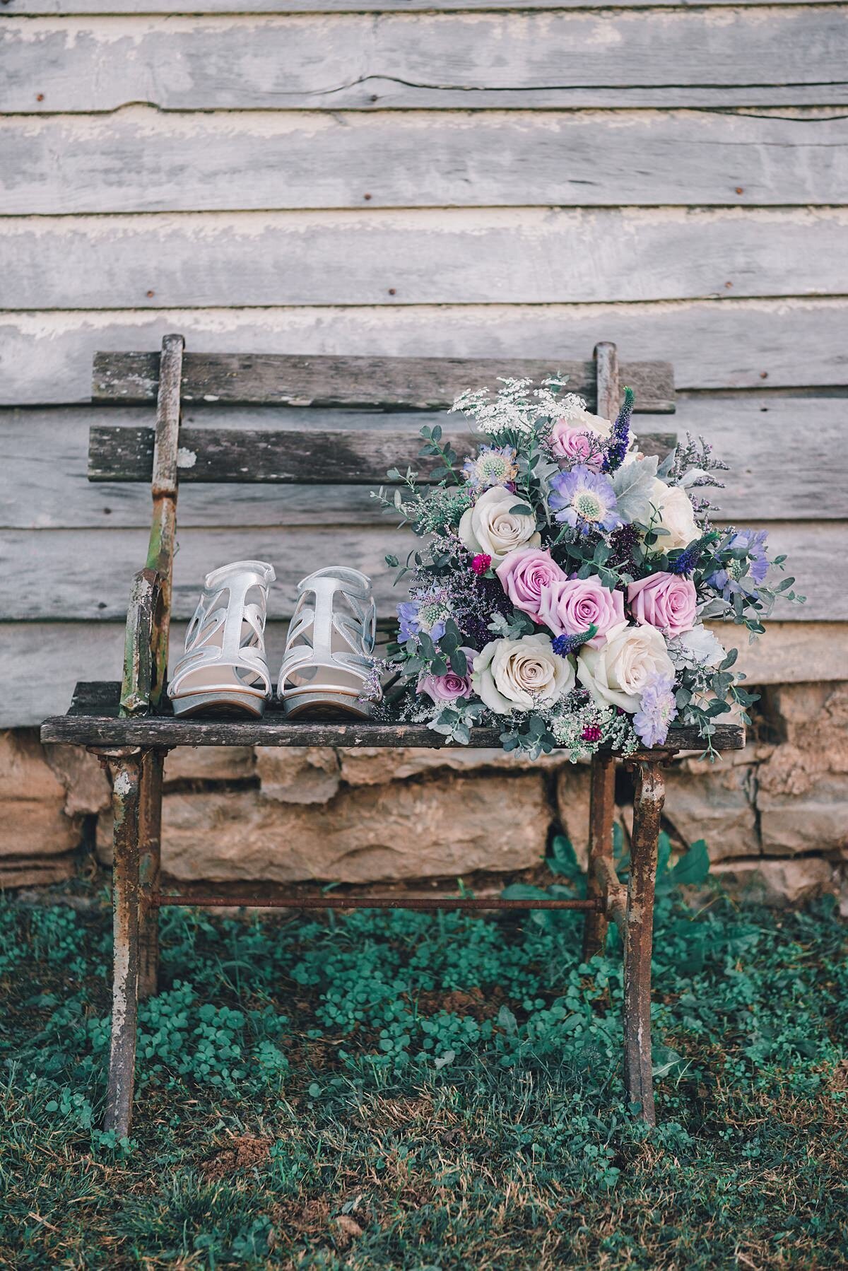 A pair of strappy wedding shoes sitting on a rustic barn wood bench next to a large bridal bouquet  of purple flowers, pink roses, ivory roses, white roses and white queen annes lace. with hints of greenery made by a Murfreesboro florist.