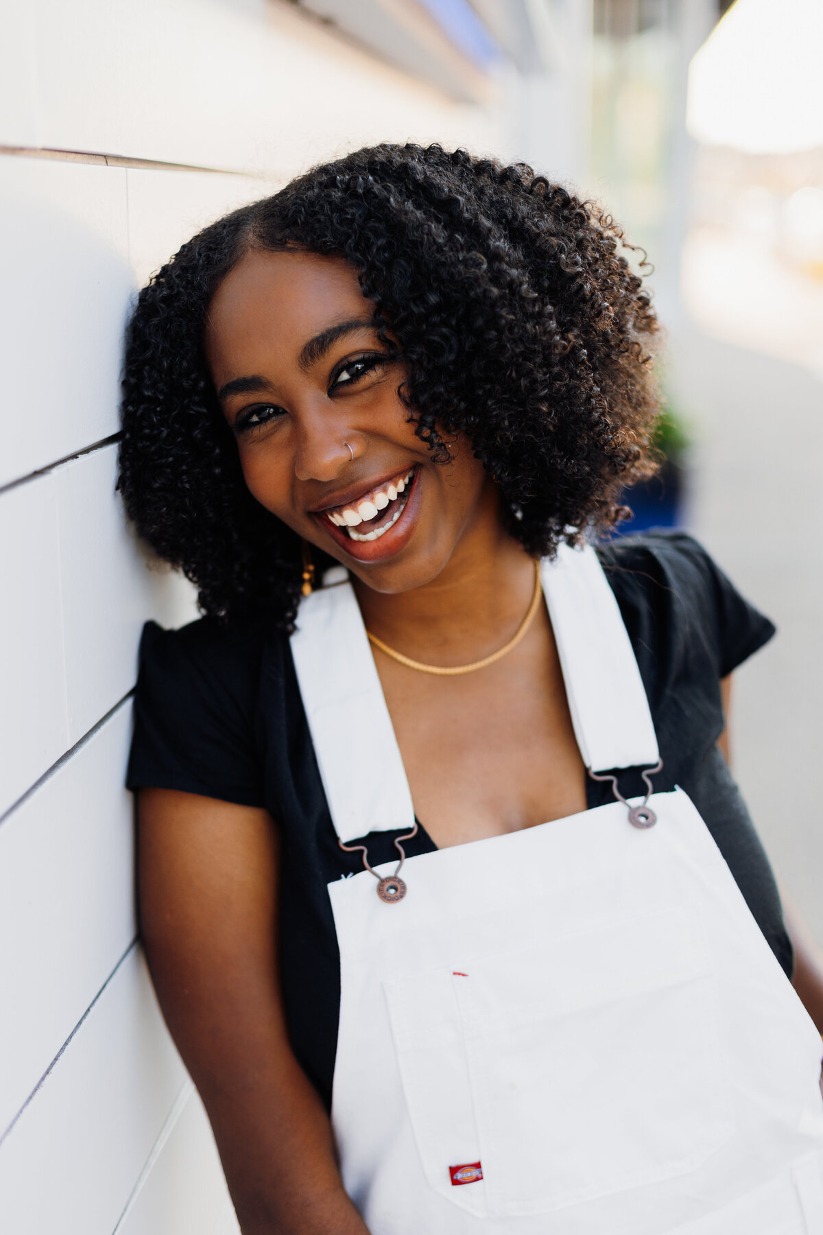 Senior girl in a black top and white overalls leaning against a white wall