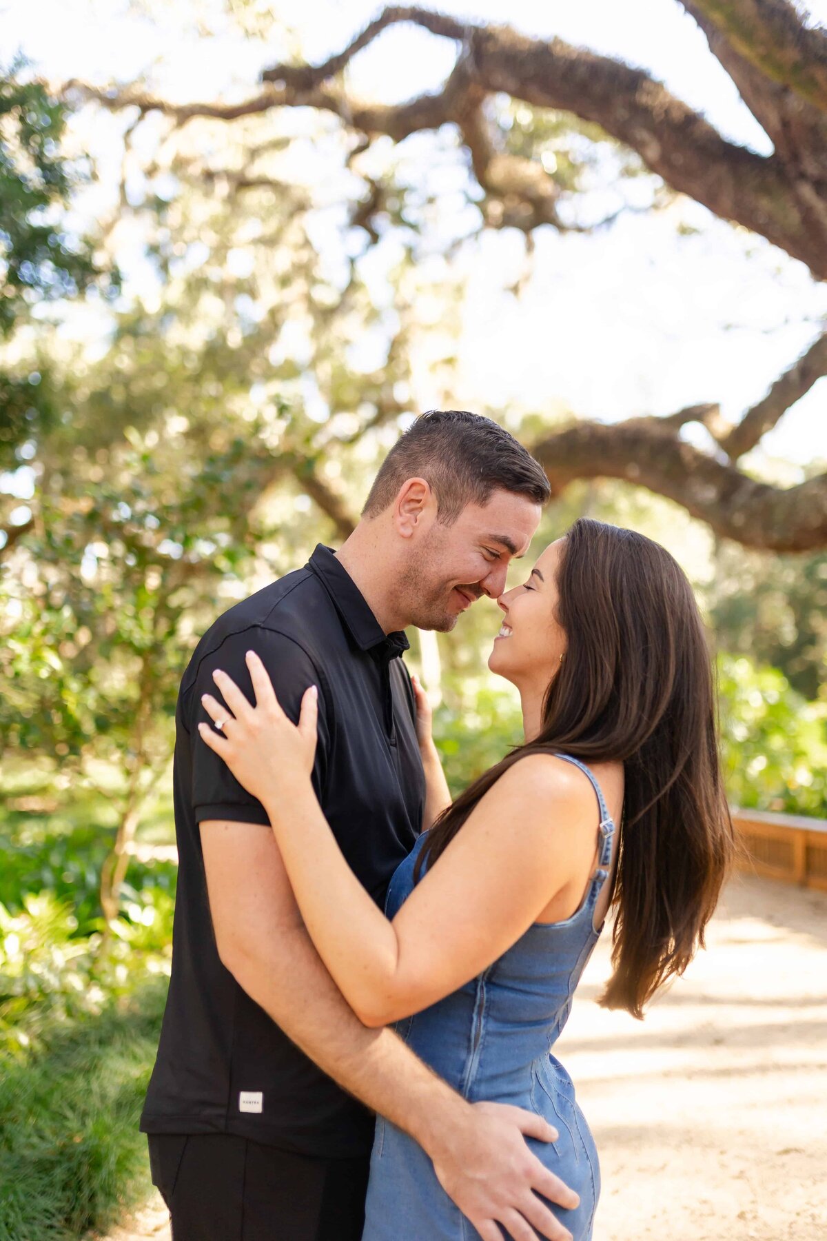 A couple shares a joyful, intimate moment as they lean in for a forehead-to-forehead embrace beneath sprawling oak trees in Jacksonville, Florida. The bride-to-be, in a denim dress, holds her fiancé close, both smiling with their eyes closed in laughter. This candid, sunlit portrait captures the natural chemistry and happiness of their engagement. Captured by Heartful Films, a Jacksonville engagement photographer and videographer specializing in relaxed, true-to-color storytelling.