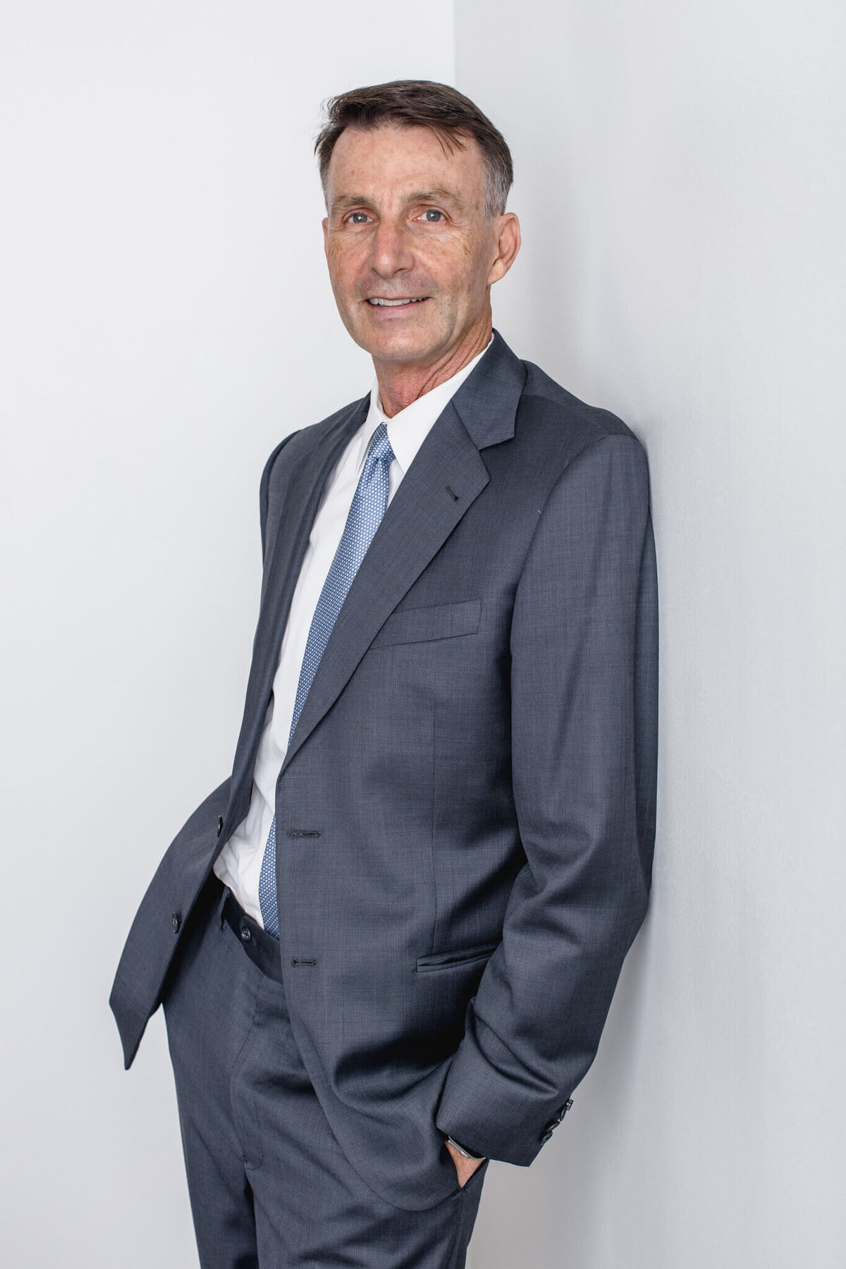 An older man with dark hair wearing a dark grey suit jacket and dress pants with his hands in his pockets, leaning his left shoulder onto a white wall and smiling at the camera in a studio in Tustin.