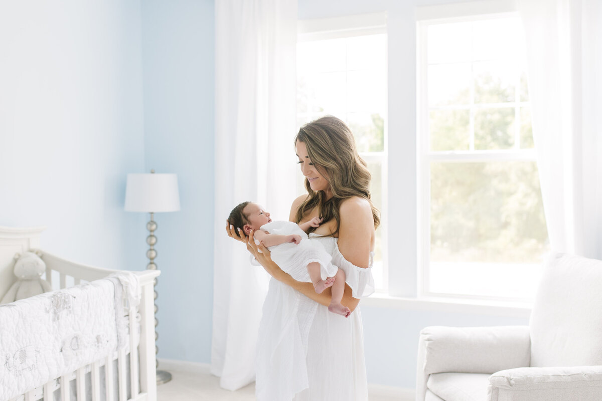 mother holding newborn in front of window  in light filled white and blue nursery