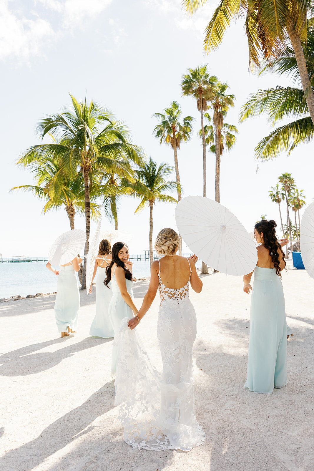 A bridal party moment with the bridesmaids in pastel blue dresses standing on the beach with white parasols.