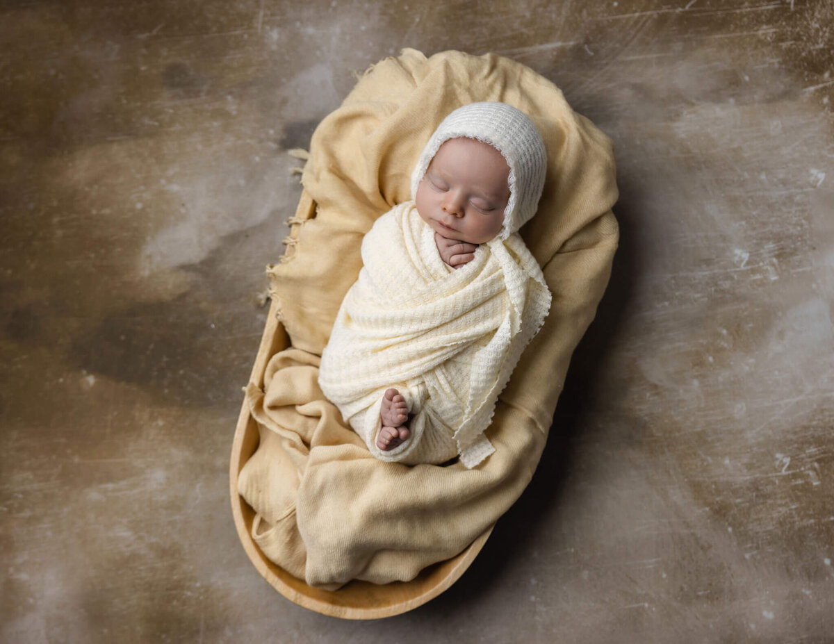 newborn baby wrapped in cream fabric wearing a cream bonnet asleep in a wooden bowl