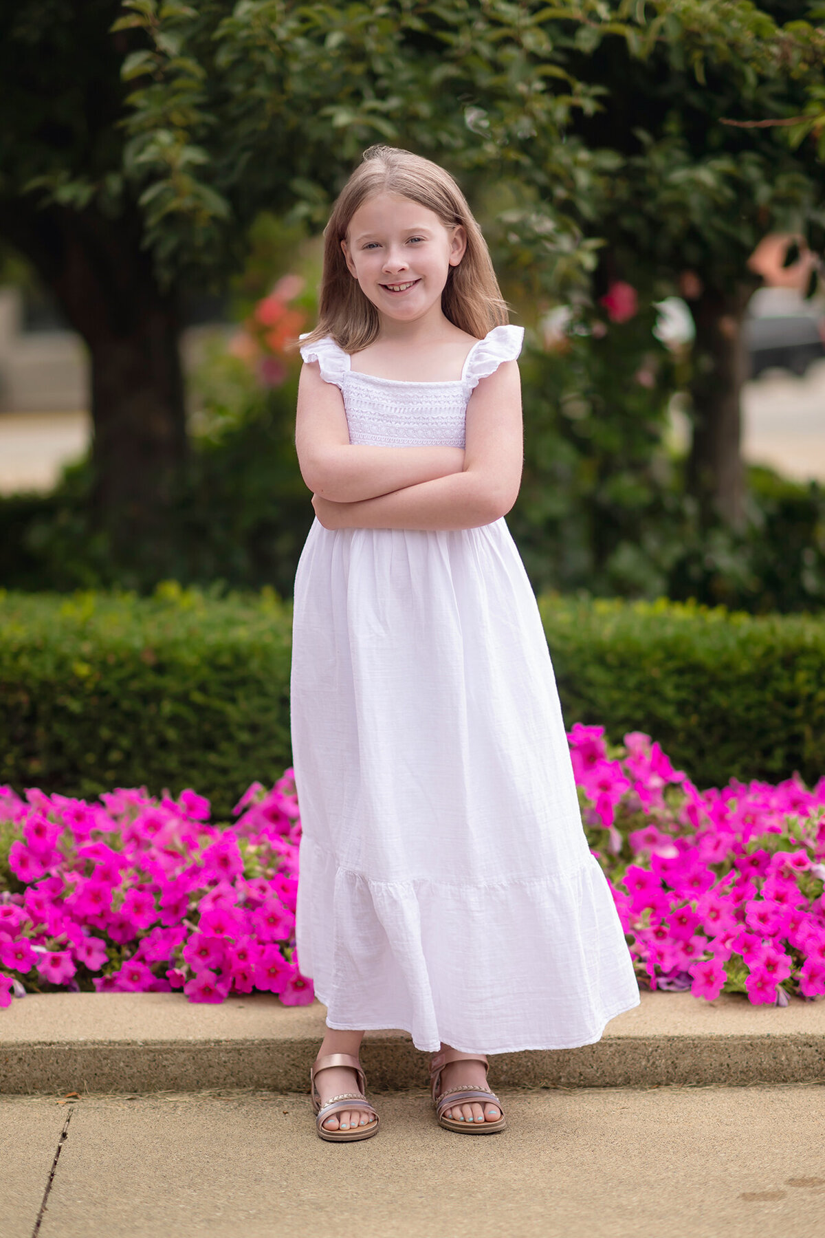 A young girl stands in her white dress in front of flowers