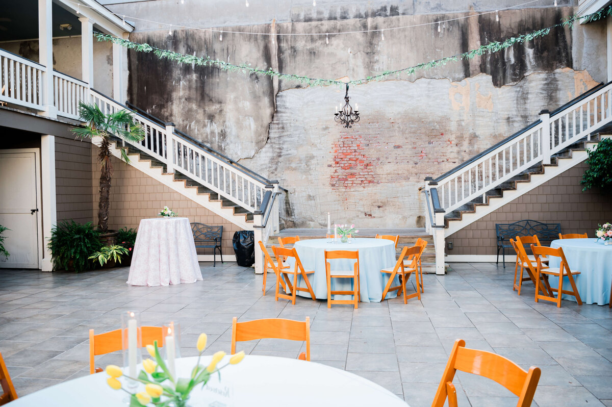 A bright courtyard set up for an event features round tables with light blue tablecloths, wooden chairs, potted plants, and string lights hanging above.