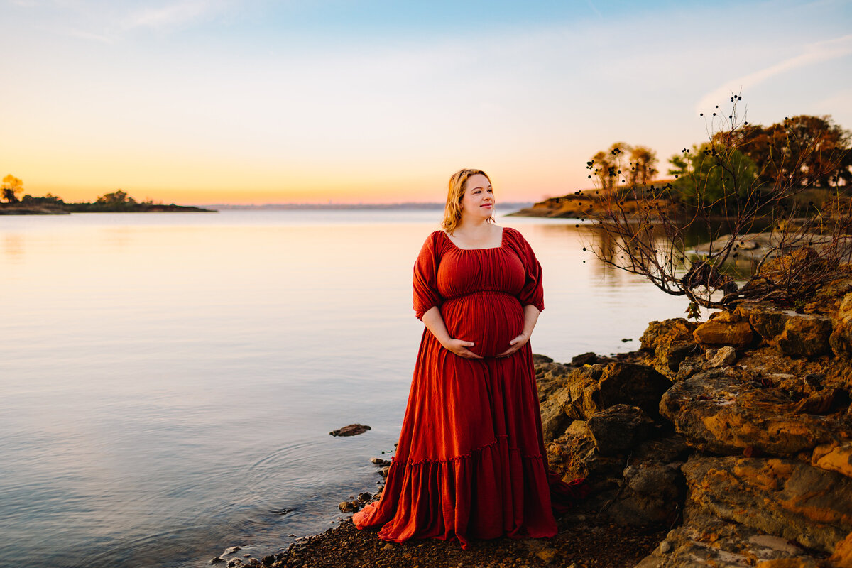 A stunning maternity photo of a woman dressed in a long red dress, standing gracefully in front of a serene lake. The red dress contrasts beautifully with the tranquil waters and surrounding nature