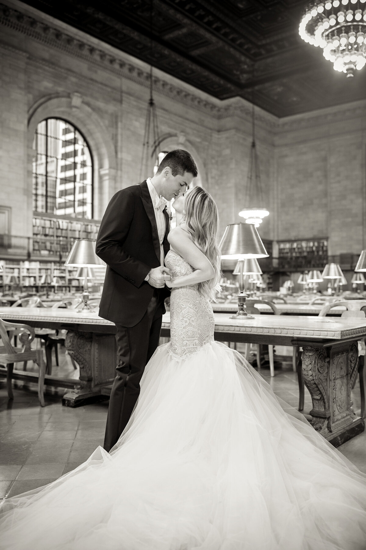 The photo captures a couple in wedding attire sharing an intimate moment at the iconic New York Public Library, beautifully captured by the talented photographer, Christian Oth. East West Films, videographers based in New York, also expertly captured this special moment. The bride, dressed in a flowing gown, and the groom, in a sharp suit, stand closely together amidst the grandeur of the library's elegant chandeliers, towering shelves of books, and rows of study tables adorned with ornate lamps. The entire scene is portrayed in classic black and white, adding a timeless and romantic quality to the moment.